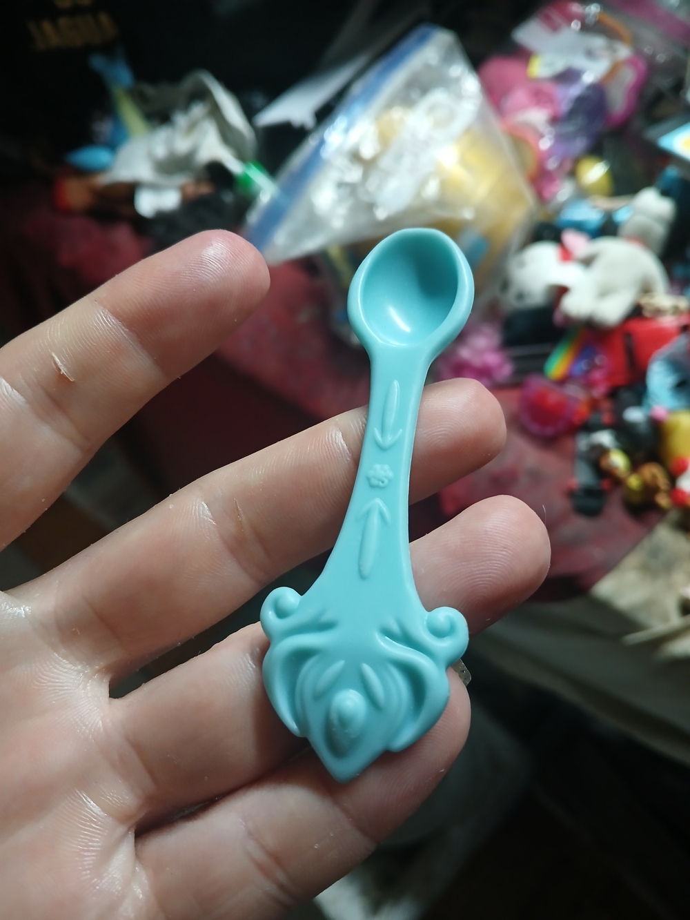 light blue or teal plastic toy spoon with an ornate, scrolled handle design.